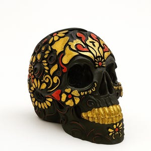 Peut inclure: Crâne décoratif noir avec des motifs floraux dorés et rouges. Le crâne présente des détails dorés sur les dents et autour des yeux. Un objet décoratif sur le thème du Jour des Morts ou d'Halloween.