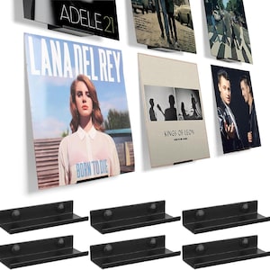 Könnte beinhalten: Eine Sammlung von Albumcovern, darunter Lana Del Reys "Born to Die" und Kings of Leon's "When You See Yourself", präsentiert auf schwarzen Wandregalen. Die Albumcover zeigen verschiedene Farben, Texte und Bilder.