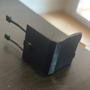 May include: A black, angular device with two attached arms and connectors. The device has a layered construction, with visible lines indicating its 3D-printed origin. The object is positioned on a dark wooden surface.