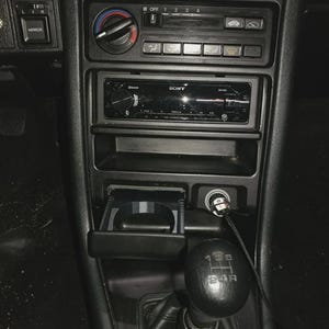 Könnte beinhalten: Die innere Mittelkonsole eines Fahrzeugs verfügt über ein schwarzes Armaturenbrett mit einem Sony-Autoradio, Klimareglern, einem ausziehbaren Getränkehalter, einer 12-Volt-Steckdose mit Ladekabel und einem manuellen Schaltknauf mit 5-Gang-Schema.