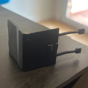 Puede incluir: Un objeto rectangular negro con dos brazos que se extienden desde los lados. Cada brazo termina en una pequeña forma cuadrada. El objeto está sobre una superficie de madera marrón oscuro.