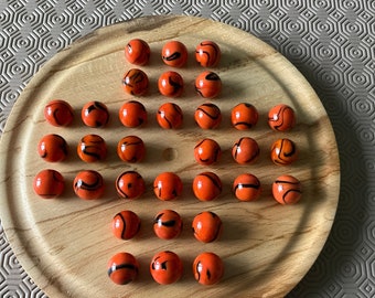 Puede incluir: Un tablero de juego de madera con 24 canicas naranjas con remolinos negros. Las canicas están dispuestas en un patrón circular en el tablero.
