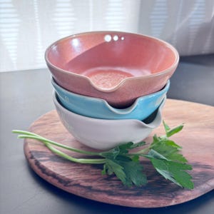 May include: A stack of three ceramic bowls with a spout, featuring a coral pink top bowl, a light blue middle bowl, and a white bottom bowl. The bowls are smooth with a glossy finish and are arranged vertically on a wooden surface.