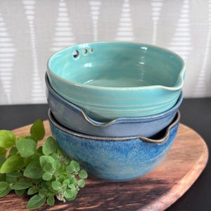 Peut inclure: Trois bols en céramique empilés, dans des tons de bleu et turquoise. Le bol supérieur comporte trois petits trous. Chaque bol a un bec verseur. Ils reposent sur une planche en bois. Des herbes vertes fraîches sont au premier plan.