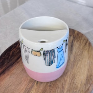 May include: A white ceramic toothbrush holder with a pink base. The holder features a hand-drawn illustration of clothes hanging on a washing line, including a blue dress and striped shirt. A small hole is at the top.