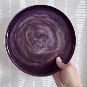 May include: A circular ceramic plate with a deep purple glaze and a white rim. The plate's centre has a swirling pattern of purples and pinks. The plate is held by a hand, showcasing its size and design. This decorative plate is suitable for serving food.