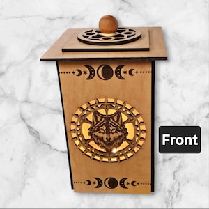 May include: Wooden box with a wolf design and moon phases on the front. The box has a wooden lid with a circular design on top. The box is lit from inside, illuminating the wolf design.