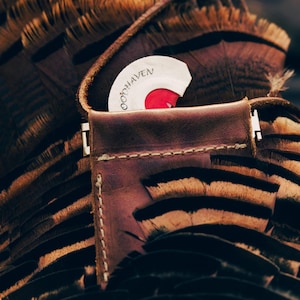 May include: Close-up of a brown leather wallet with a white and red circular object inside. The wallet is stitched with white thread and has a metal clasp. The background features brown and black feathers.