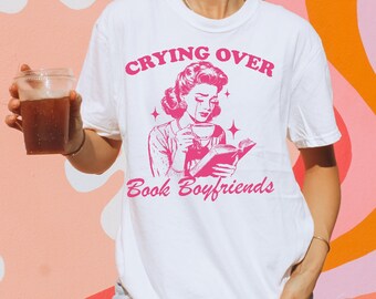 Op de afbeelding: Wit T-shirt met roze tekst die "Crying Over Book Boyfriends" luidt. De afbeelding toont een vrouw in een vintage jurk die een kopje koffie vasthoudt en een boek leest.