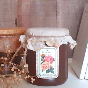 May include: Two glass jars containing brown liquid, both with white fabric lids. The jar on the right has a label that reads "Margaret" and features a floral design. The jars are positioned on a wooden surface with a stack of books behind them.
