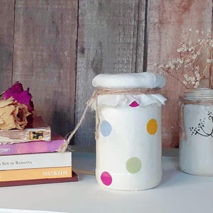 May include: Two white ceramic jars with decorative designs. The jar on the left has a white fabric lid and is decorated with colourful polka dots. The jar on the right has a simple black floral design. Both jars are tied with twine.
