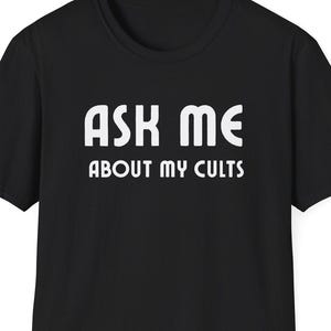 May include: Black t-shirt with the white text "ASK ME ABOUT MY CULTS". The shirt has a crew neck and short sleeves. The text is centered on the front of the shirt.
