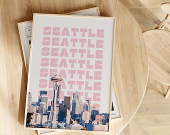 May include: A framed print with a cityscape of Seattle, Washington. The print features the Space Needle and the city skyline. The word "Seattle" is repeated in pink letters on a white background.