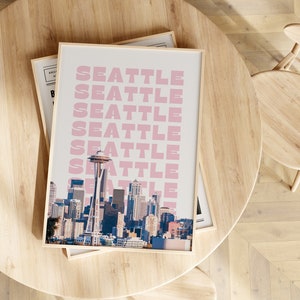 May include: A framed print with a cityscape of Seattle, Washington. The print features the Space Needle and the city skyline. The word "Seattle" is repeated in pink letters on a white background.