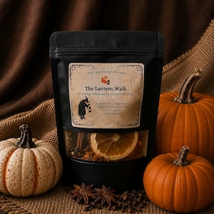 Op de afbeelding: Een zwarte zak met het label Ivy Lynne's Apothecary The Lantern Walk Nostalgic Halloween Simmer Pot Kit, met gedroogde sinaasappelschijfjes, kaneelstokjes, kruidnagels, steranijs en pepermunt, omringd door drie pompoenen.