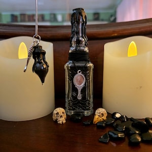 May include: A clear glass bottle topped with black wax drips, featuring a silver-toned hand mirror pendant on a black cord. A silver pendulum with a black wax-dipped charm hangs nearby, accompanied by two small skulls and black stones.