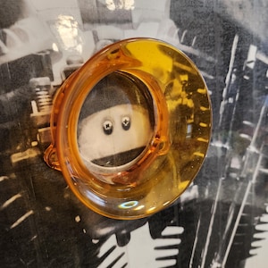 Op de afbeelding: Een transparant, amberkleurig glazen object met een cirkelvorm en een klein, bevestigd handvat. Het object staat tegen een zwart-witte achtergrond met een motorfietsmotordesign.