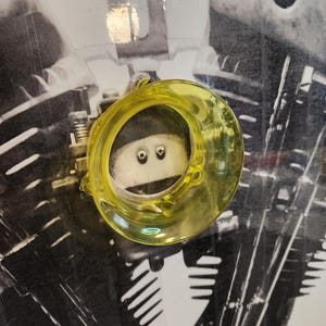 Op de afbeelding: Een geel glazen ornament met een cirkelvorm. Het transparante ornament reflecteert licht. De achtergrond is een zwart-wit afbeelding van een motorfietsmotor.