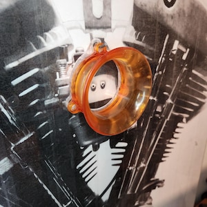 Op de afbeelding: Oranje plastic motorfiets luchtinlaatfilter. Het filter is rond en heeft een glad oppervlak. Het is bevestigd aan een metalen beugel met twee schroeven.