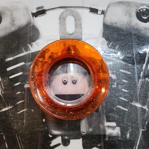 Op de afbeelding: Een oranje, cirkelvormige motorfiets richtingaanwijzer met glitteraccenten. De richtingaanwijzer is gemonteerd op een zwart-wit afbeelding van een motorfietsmotor.