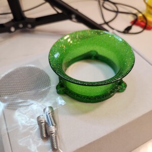 Op de afbeelding: Een groen, met glitter doordrenkt trechtervormig object met een brede opening en een cirkelvormige basis. De afbeelding toont ook een klein metalen gaasscherm en verschillende zilveren schroeven. De trechter heeft een decoratief ontwerp.