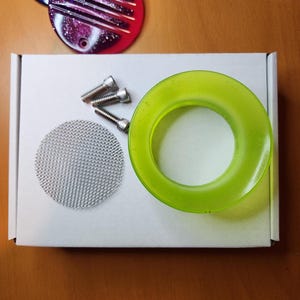 Op de afbeelding: Een witte doos met een groene siliconen ring, een zilveren gaaszeef en vier zilveren schroeven. De doos staat op een houten oppervlak.