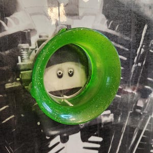 Op de afbeelding: Een doorschijnend groen, cirkelvormig object met witte spikkels. Het object staat tegen een zwart-witte achtergrond. Het object heeft een gat in het midden, dat een cartoongezicht onthult.