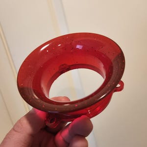 Op de afbeelding: Een transparant rood trechtervormig object met ingebedde glitter. Het object heeft een brede, cirkelvormige opening en een kleinere, cirkelvormige uitlaat. De trechter wordt in een hand gehouden, met een witte deur op de achtergrond.