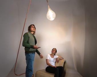 May include: A tall, arched floor lamp featuring a thin, curved copper-colored metal stand and a single, teardrop-shaped glass pendant light that emits a soft, warm glow. The lamp base rests on the floor, extending upward in a long, slender arc.