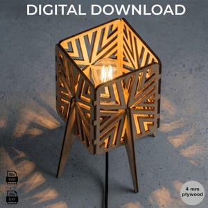 Może przedstawiać: Kwadratowa drewniana lampa z geometrycznym wzorem wyciętym laserowo i żarówką w środku. Widoczny jest tekst DIGITAL DOWNLOAD, SVG, DXF oraz 4 mm plywood. Lampa ma cztery nogi i emituje ciepłe światło, rzucając skomplikowane cienie na powierzchnię.