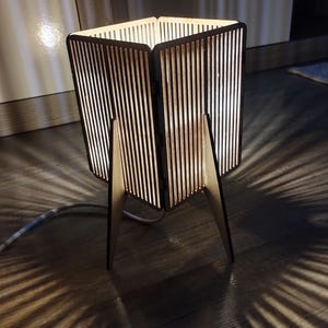 May include: A wooden table lamp with a geometric design. The lamp has a square base and a rectangular shade with vertical slats. The lamp is lit and casting a warm glow.