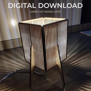 Könnte beinhalten: Eine lasergeschnittene Stimmungsleuchte mit einem einzigartigen Design. Die Lampe hat einen rechteckigen Schirm mit vertikalen Lamellen, der von drei Holzbeinen getragen wird. Der Text "DIGITAL DOWNLOAD" und "LASER CUT MOOD LIGHT" ist sichtbar. Die Lampe wirft einen Schatten.