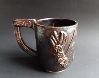 May include: A handcrafted ceramic mug with a dark brown glaze. The mug features a detailed handle and embossed relief designs on the body, including a stylized wing motif. The mug is approximately 4 inches tall and suitable for hot beverages.