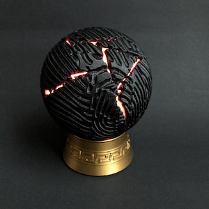 Può includere: Un oggetto sferico con una superficie nera testurizzata caratterizzata da profonde crepe irregolari che emettono una luce rossa brillante. La sfera poggia su una base circolare color oro metallizzato decorata con un motivo geometrico greco.