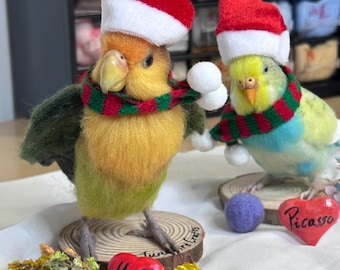 Puede incluir: Dos figuras de aves de fieltro con gorros de Papá Noel rojos y blancos y bufandas a rayas rojas y verdes. Un ave es naranja y verde, la otra amarilla y azul. Cada una sobre una rodaja de madera con un corazón rojo etiquetado Yuko o Picasso.