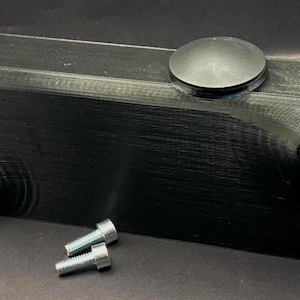Puede incluir: Un objeto rectangular negro con una parte superior redondeada y dos accesorios dorados. Una tapa circular negra está en la parte superior. Dos pernos plateados están frente al objeto. El objeto parece estar impreso en 3D.
