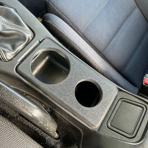 Könnte beinhalten: Schwarze Kunststoff-Autokonsole mit zwei kreisförmigen Getränkehaltern und einem rechteckigen Ablagefach. Die Konsole ist in den Innenraum des Autos integriert, mit einem schwarzen Schalthebel und einem grauen Stoffsitz im Hintergrund.