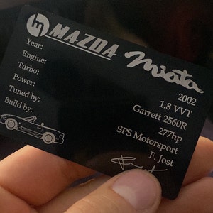 Könnte beinhalten: Eine schwarze Metallplakette mit den Worten "MAZDA Miata" und Informationen über einen Mazda Miata aus dem Jahr 2002, einschließlich Hubraum, Turbolader, Leistung und den Namen des Tuners und des Herstellers.