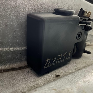 Może przedstawiać: Czarny element samochodowy z cylindryczną nasadką i srebrnym napisem "カッコイイ Kakkoi Racing Dept.". Element ma prostokątny korpus i jest zamontowany na metalowej powierzchni.