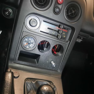 Puede incluir: Un interior de coche negro con un medidor montado en la consola central. El medidor tiene una cara blanca y números negros. La consola también tiene algunas ranuras vacías para medidores adicionales.