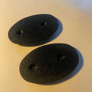 Könnte beinhalten: Zwei schwarze, ovale Kunststoffteile mit jeweils zwei Löchern. Die Teile haben eine strukturierte Oberfläche.