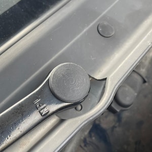 Può includere: Primo piano di un fermo per cofano di un'auto in metallo nero con una maniglia circolare in plastica nera. La maniglia ha una superficie testurizzata e il fermo è etichettato "N.F.I."