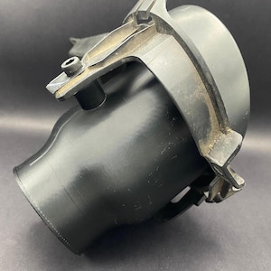 Può includere: Un componente automobilistico in plastica nera con un corpo curvo e cilindrico e una struttura a staffa. Il componente ha una finitura opaca e sembra essere utilizzato nel motore o nell'impianto di scarico di un veicolo. La staffa ha una vite metallica.