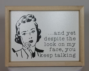 May include: A framed print with a black and white illustration of a woman with a thoughtful expression. The text reads "...and yet despite the look on my face, you keep talking."