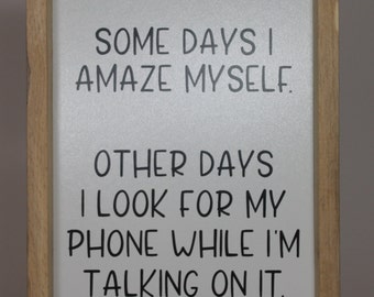 May include: A framed sign with a white background and black text that reads "Some days I amaze myself. Other days I look for my phone while I'm talking on it."