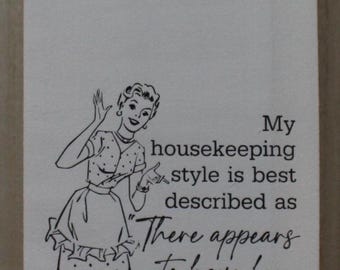 May include: A white tea towel with a black and white illustration of a woman in a polka dot dress. The text reads "My housekeeping style is best described as "There appears to have been a struggle."