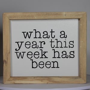 May include: A wooden framed sign with white background and black text that reads "what a year this week has been".