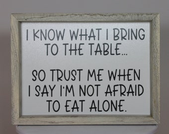 May include: A rectangular wooden sign with a distressed, light gray frame. The white background features black text that reads, "I KNOW WHAT I BRING TO THE TABLE... SO TRUST ME WHEN I SAY I'M NOT AFRAID TO EAT ALONE."
