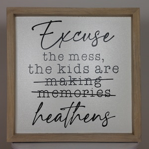 May include: A framed sign with the text "Excuse the mess, the kids are making memories heathens" in black lettering on a silver background.