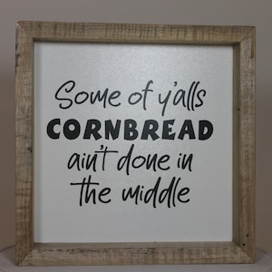 May include: A framed wood sign with white background and black text that reads "Some of y'alls cornbread ain't done in the middle."